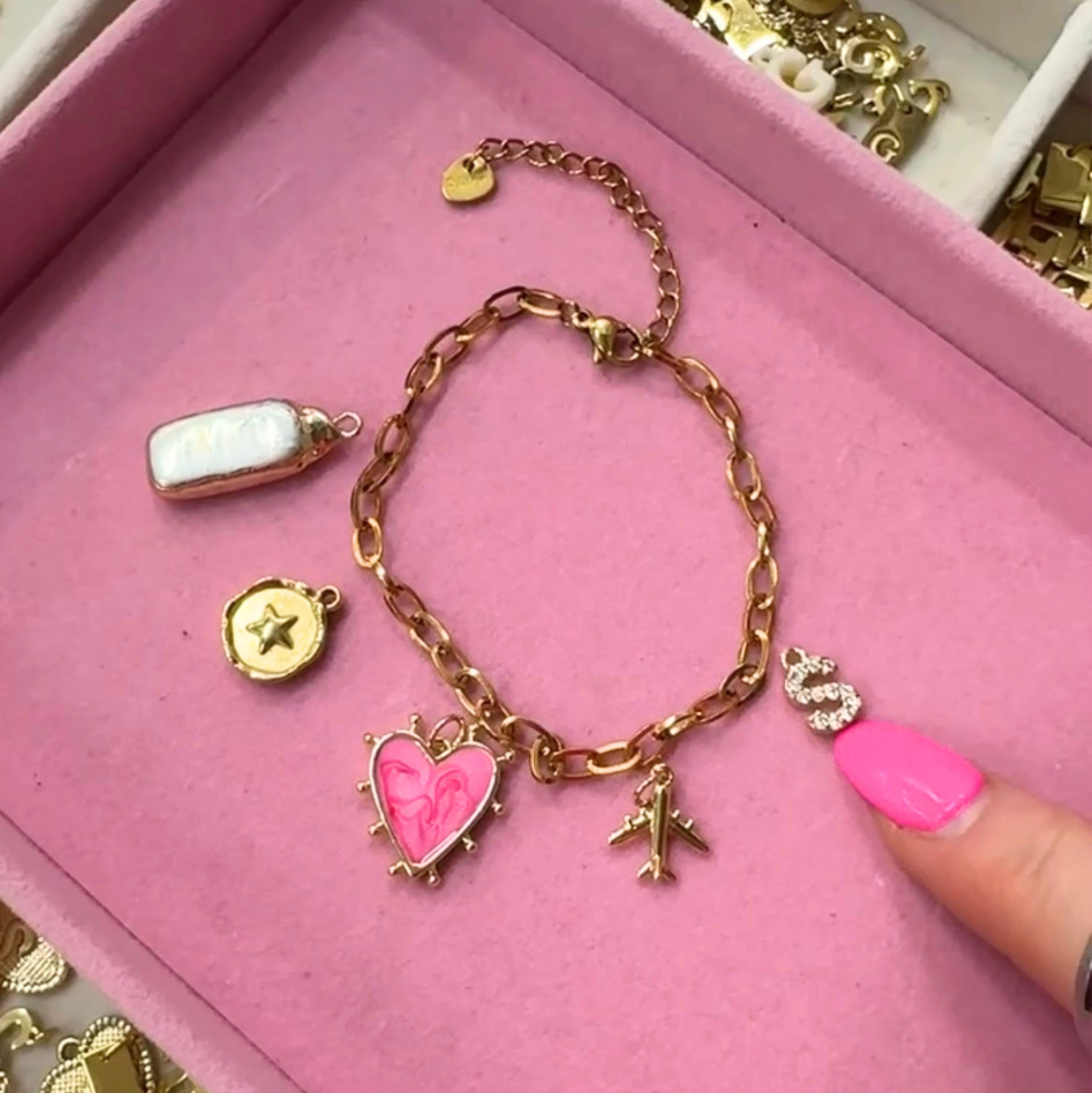 A gold-tone charm bracelet with a pink heart charm, airplane charm, and small letter charms, displayed on a pink jewelry box next to a finger with pink nail polish.