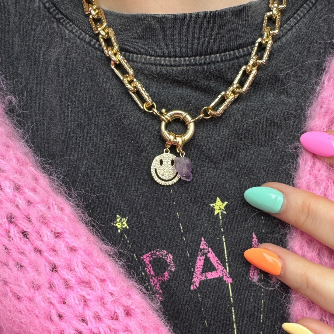 A close-up of a gold chain necklace with a smiley face charm and a small purple heart pendant, worn over a dark gray graphic t-shirt and a pink knit sweater. A hand with pastel-colored nails is visible on the right side.