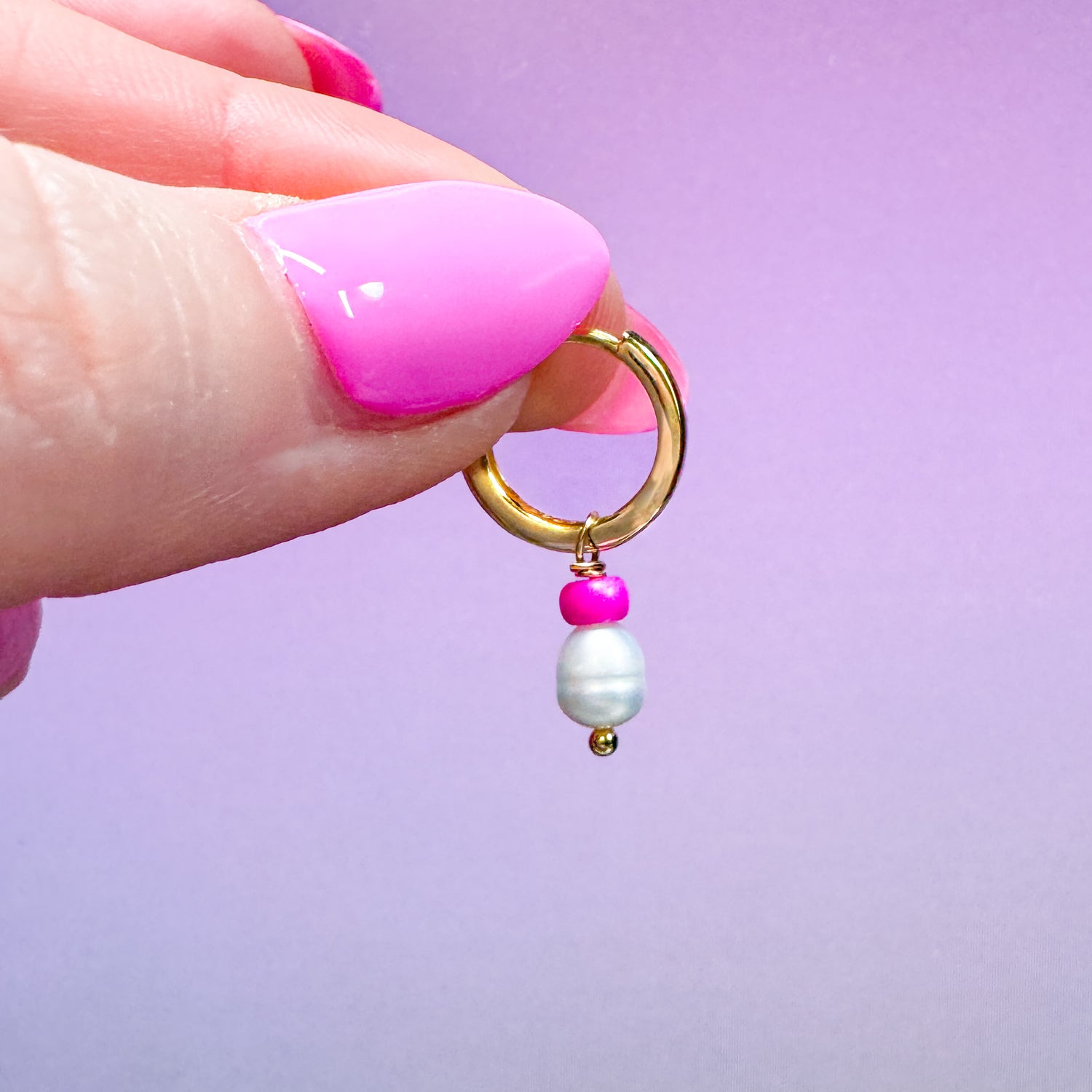 Gold hoop earring with pink and white beads held by a hand against a purple background