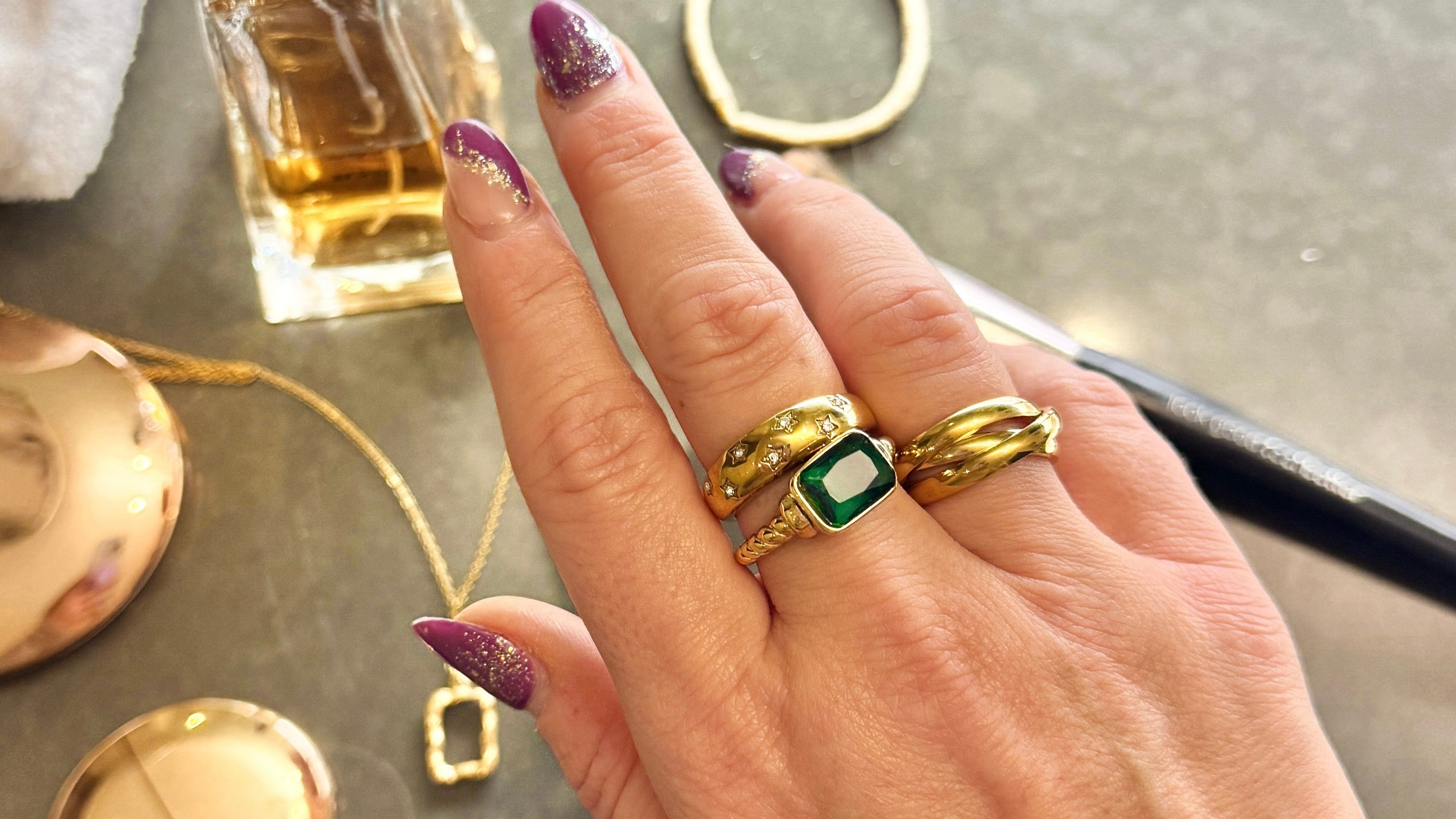 Hand wearing gold rings with a small green gemstone, with jewelry and a perfume bottle in the background.