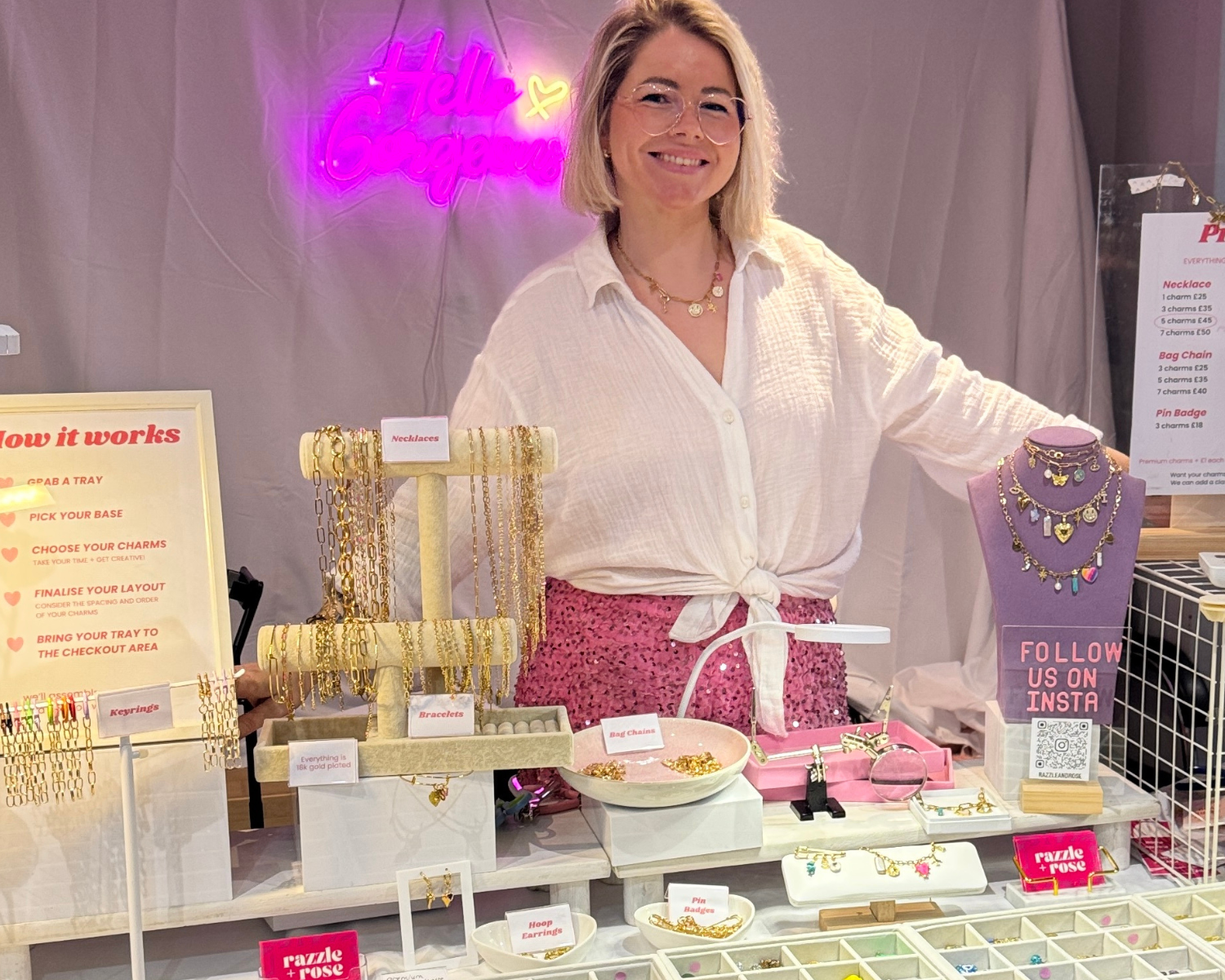 A woman stands behind a jewelry display booth at what appears to be a fair or market, wearing a white button-up shirt and pink patterned skirt. The booth is filled with necklaces, bracelets, and rings on display stands, with a pink neon sign in the background.