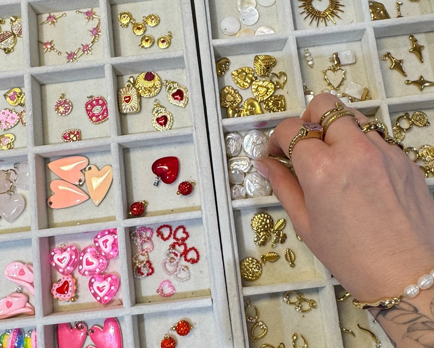 A hand with rings and a bracelet reaches into a tray of assorted jewelry findings, including gold-toned heart charms, silver heart charms, and pink enamel heart studs, next to another tray of earrings.